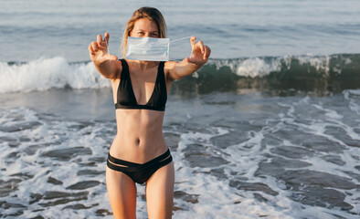 Happy girl in bikini, with medical protective mask on her hands, walking by the sea. Summer, coronavirus, quarantine. Covid-2019. Virus stop.