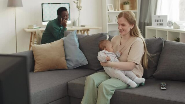 Medium shot of Caucasian woman sitting on couch at home and nursing her infant daughter while African man having cellphone call on background