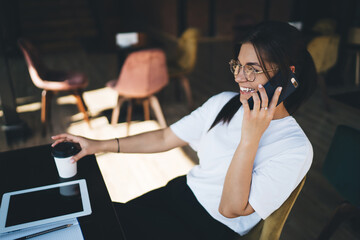 Cheerful young caucasian female ein eyewear talking on mobile phone sitting on coffee break, smiling woman 20s in trendy eyewear making smartphone call satisfied with connection and tariff