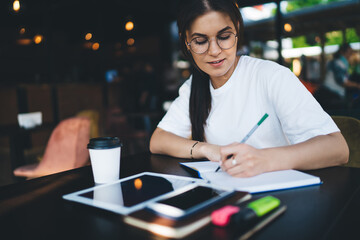 Creative caucasian female blogger using notebook for sketching and making notes sitting in cafe interior with digital tablet, clever positive beautiful woman 20s doing homework using touchpad