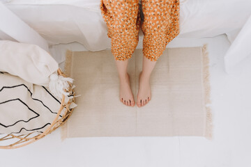 Young woman sitting on the bed put her feet on a rug made of natural materials. Eco-friendly...