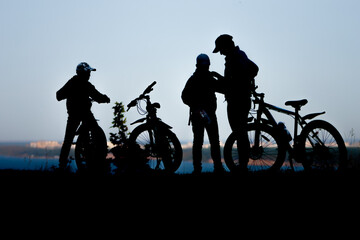 Obraz premium Silhouettes of figures of a father with two sons on a Bicycle ride in the Park by the river. The concept of a healthy lifestyle in the family.