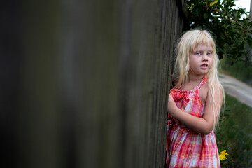 Little girl in the village. White hair, pink dress. Wooden fence in the background.