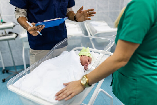 Young Doctor And Nurse Working In Maternity Ward. They Are Collecting Data, Measuring Weight And Wrapping The Newborn Baby.