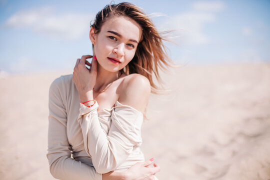 A young, slender girl in a beige dress poses in the wind in the desert