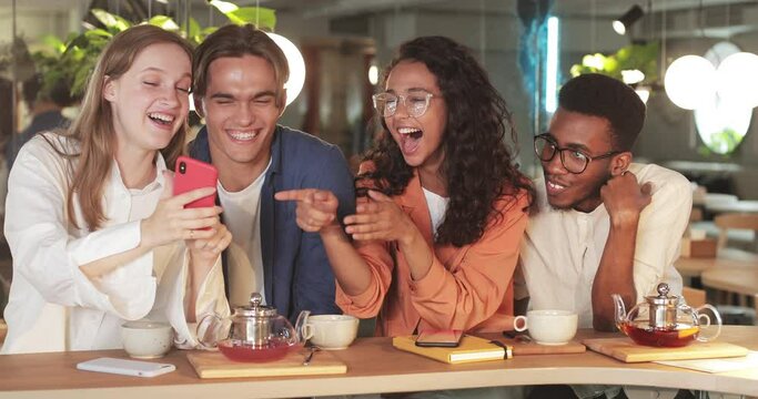 Crop view of coworkers looking at phone screen while spending good time during lunch. Millennial smiling friends viewing photos on smartphone while sitting at table in modern cafe.