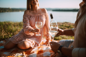 Close - up of A cute blonde woman in a pastel pink dress and a man drinking white wine on the beach at sunset. A loving couple celebrating and enjoying a picnic on the river bank