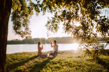 A cute blonde woman in a pastel pink dress and a caucasian man drinking white wine on the beach at sunset. A loving couple celebrating and enjoying a picnic on the river bank