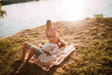An attractive handsome blonde woman and a caucasian man lying in her lap on the beach at sunset, eating grapes and drinking wine. A loving couple on a picnic by the river