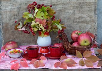 On a pink napkin are two cups of coffee, a plate with candied fruits, an original basket with apples, a bouquet with viburnum leaves and berries. Red autumn leaves lie nearby.