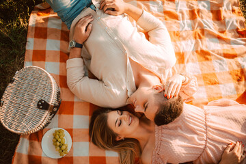 Top view of a caucasian man and a blonde woman  lying on an orange blanket at a picnic,  and next to them is a basket of food and grapes