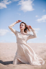 Fototapeta na wymiar A young, slender girl in a beige dress poses in the wind in the desert