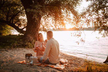 A beautiful caucasian woman in a pastel pink dress and a man are sitting on the beach and eating a cake. A loving couple celebrating a birthday on the river bank
