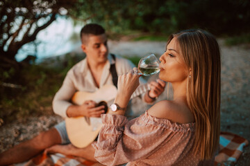 A beautiful caucasian woman in a pastel pink dress  drinking wine and a man playing guitar are sitting on the beach. A loving couple on a picnic by the river