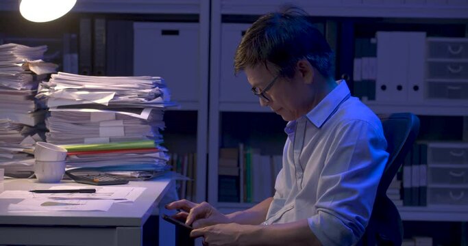 Senior asian business man is sitting at desk covered with stack of paperwork and using smart phone. Alone male relax using mobile browsing internet, social networks while working hard at late night.