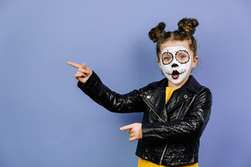 Portrait of cute little girl with bright scary makeup, dressed in black leather jacket, points aside on purple studio copy space, shows place of Halloween party. Mexican culture, Day of Death concept