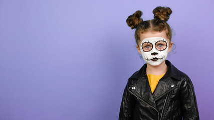 Portrait of child girl looking at camera, wears black leather jacket, isolated on purple studio background with copy space. Little girl wears frightening makeup. Kid prepares for mexican day of dead