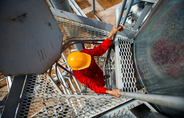 Oil refinery worker in red work wear and yellow helmet. Grey metal grid floor and stairs. Distillation column on refining complex (plant). CNPC company.
