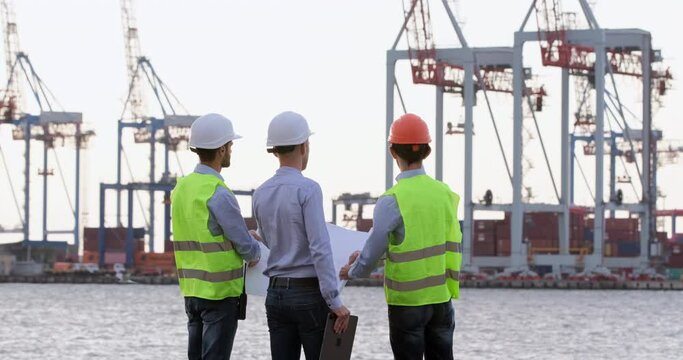 Port workers in special clothes discuss the drawing on white paper against the background of the seaport. The contractor gives instructions to subordinates.