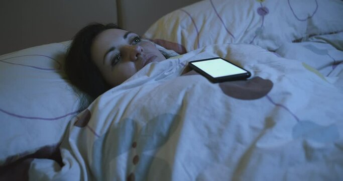 Sad lonely woman ignores calls. A pensive woman, with an apathetic look, lying on the bed, wrapped in a blanket and looking at the ceiling. Night. Phone calling, woman ignoring calls. Seperation 