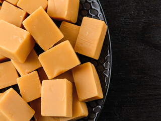 Top view of caramel vanilla fudge candies on a black textured ceramic plate over dark wood table. Fresh tasty chewy bonbons made of milk and sugar. Square pieces of delicious soft sweets.