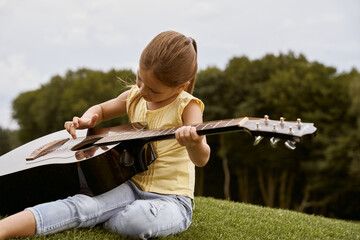 Cute little girl in casual clothes sitting on a green grass in park and learning how to play guitar