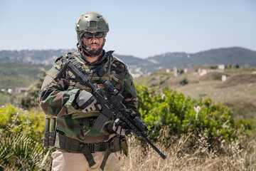 Fototapeta premium Private military company mercenary, brutal looking special forces fighter in battle uniform and plate carrier, wearing radio headset and sunglasses, holding service rifle in hands, ready to fight