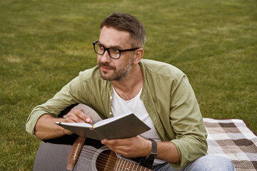 Young thoughtful man wearing eyeglasses holding acoustic guitar and composing a song while sitting on a green grass outdoors