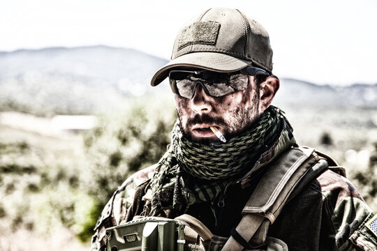Shoulder portrait of commando fighter, professional mercenary or special forces shooter with dirty face, wearing shemagh scarf, ballistic glasses and cap, carrying service rifle and smoking cigarette