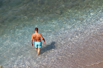 Man in blue shorts going to swim in a sea. Aerial view to the pebble beach with transparent water, background for vacation and travel