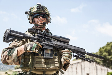 Fototapeta premium Half-length portrait of special forces sniper or marksman, army soldier in camouflage uniform and helmet with radio headset, standing on background of blue sky, holding sniper rifle with optical scope
