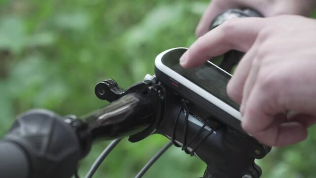 Man uses smart bicycle computer fixed on handlebars mountain bike, cyclist using navigator to find GPS coordinates while cycling in forest. Cycle computer with offline maps for tourism and travel.
