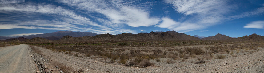 Fototapeta na wymiar Traveling along the dirt road. Panorama view of the empty route across the arid desert and mountains under a beautiful blue sky with clouds.
