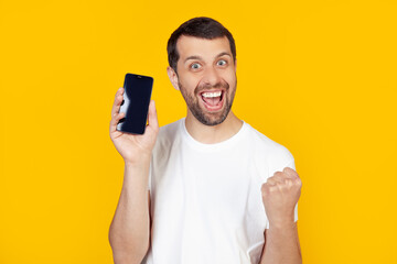 A young man with a beard in a white T-shirt shows a smartphone screen, shouts with pride and celebrates victory and success, is very excited, rejoices in emotions. Stands on isolated yellow background