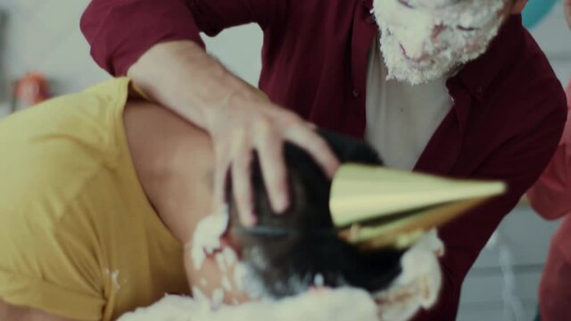 Group of funny mixed-race friends celebrating birthday. Fun guy smashing cake into his friend's face playing crazy. Party. Entertainment. Concept of humor.