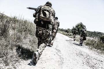Army soldiers team in combat uniform, load carriers and helmets running on countryside road, aiming...