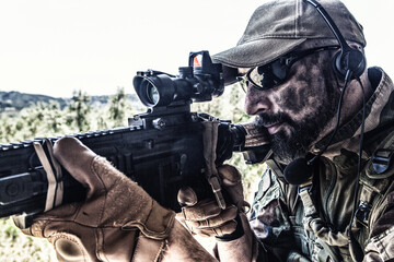 Fototapeta premium Army special forces soldier, modern combatant with dirty, unshaven face, in combat uniform, glasses and cap, equipped radio headset, aiming service rifle or light machine gun with optical sight