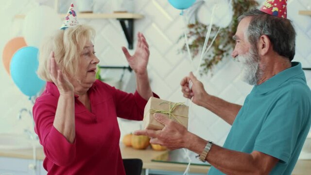 Elderly romantic man surprising wife with birthday wishes and presents at home. Happy euphoric old lady hugging her husband enjoying moments together. Holiday family.