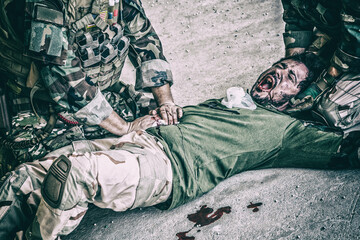 Soldiers trying to stop bleeding at wounded comrade who lying on floor, suffering and screaming in...