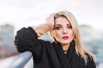 Outdoors lifestyle fashion portrait of stunning blonde girl walking on the roof. Looking at the city roofs. Thinking, enjoying view. Wearing stylish black oversized coat. Cold weather. Feeling cold