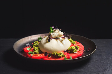 Salad of burrata cheese, tomatoes and pesto sauce in a black plate, on a dark background. Farm food and table setting concept
