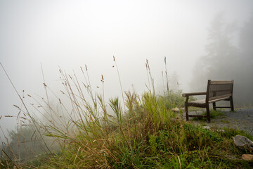 There is a dense fall fog over the mountains at the lookout where there isa bench to sit on