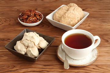 Meringue cookies with hazelnuts, almond cookies in a square bowl and pecan nuts in a small bowl, and a cup of black tea on a wooden table. Closeup