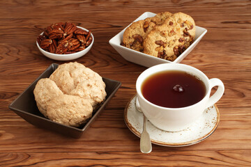 Almond cookies and oatmeal cookies with walnuts and raisins in a square bowls, and pecan nuts in a small bowl, and a cup of black tea on a wooden table. Closeup