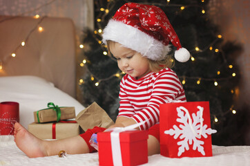 Obraz na płótnie Canvas Happy Holidays! Adorable baby girl under Christmas tree with gift boxes. Christmas present. Cure girl in Santa hat open gift box.