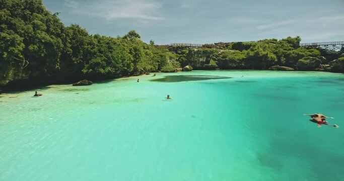 Closeup people swim at turquoise crystal clean water of Weekuri lake. Tourists landmark in green tropic forest with high bridge. Magical summer vacation in tropical paradise of Sumba Island, Indonesia