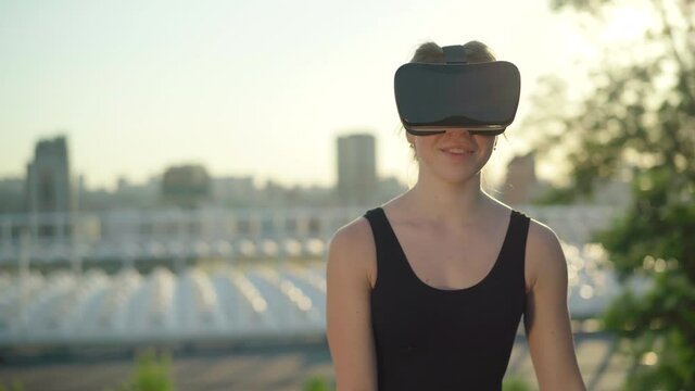Portrait of carefree Caucasian woman in VR-goggles at sunset with blurred urban city at the background. Relaxed young lady enjoying virtual reality outdoors in the evening.