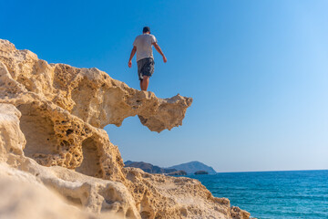 Fototapeta premium A young man perched on the rocks looking at Playa los Escullos in the natural park of Cabo de Gata, Nijar, Andalucia. Spain, Mediterranean Sea