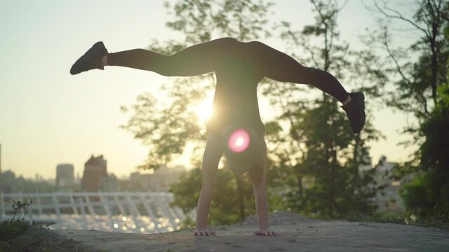 Wide shot handstand of slim young gymnast in sunrays with blurred city at the background. Legs split pose of slim flexible Caucasian woman at sunset outdoors.