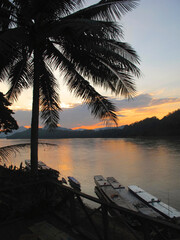 Fototapeta premium Scenic sunset view over the Mekong River in Laos, with palm trees and traditional wooden rafts and boats
